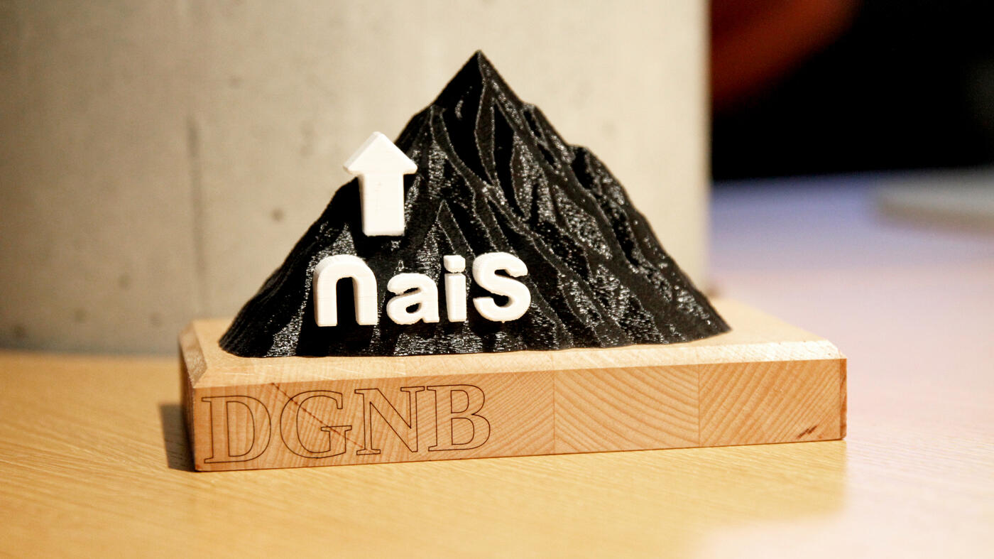 Kleine, 3D-gedruckte Bergstatuette in schwarz mit weißem NaiS-Schriftzug auf der linken Seite auf einer Holzbasis mit DGNB-Schriftzug vor grauer Betonsäule.