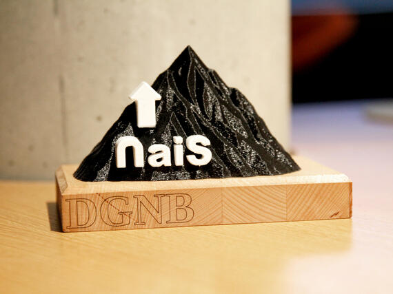 Kleine, 3D-gedruckte Bergstatuette in schwarz mit weißem NaiS-Schriftzug auf der linken Seite auf einer Holzbasis mit DGNB-Schriftzug vor grauer Betonsäule.