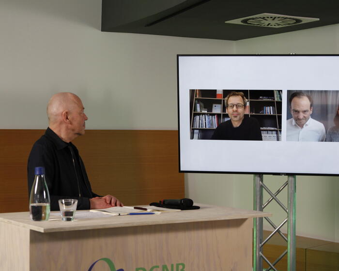 Auf dem Foto ist Prof. Amandus Samsøe Sattler hinter einem Pult zu sehen. Sein Blick richtet sich auf einen Bildschirm, auf dem zugeschaltet sind: Matthias Foitzik, Partner, foundation 5+ Architekten Ruth Klingelhöfer-Krötsch, Partnerin, Klingelhöfer Krötsch Architekten Prof. Stefan Krötsch, Partner, Klingelhöfer Krötsch Architekten