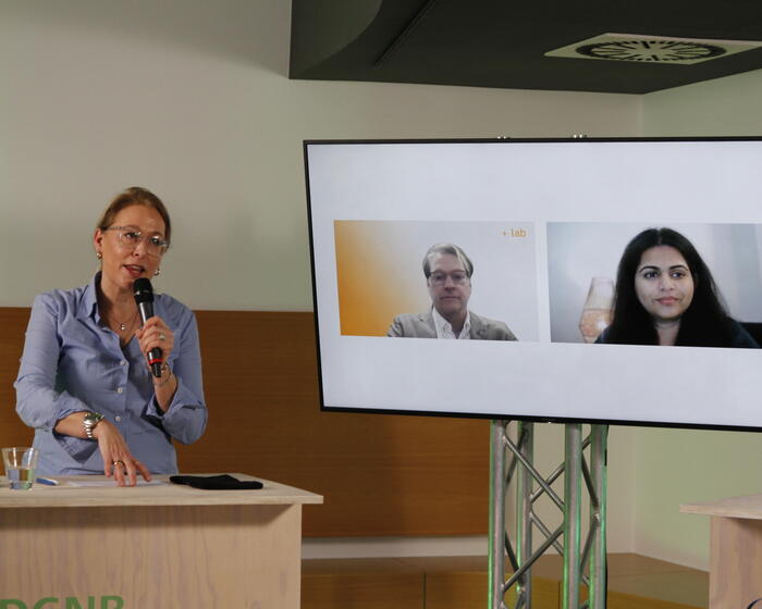 Auf dem Bild ist Dr. Christine Lemaitre mit einem Mikrophon zu sehen. Auf dem Bildschirm neben ihr sind Illya Azaroff und Rosie Paul zugeschaltet. 