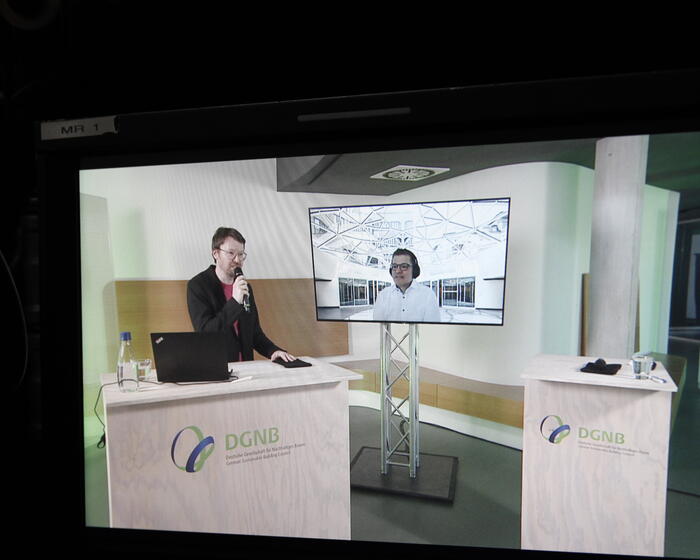 Das Foto zeigt einen Monitor. Auf diesem Monitor ist eine Studiosituation gezeit. Links: Felix Jansen (DGNB) mit Mikrofon. Rechts auf einem Monitor: Dr. Steffen Hinss, Senior Project Manager, Munich Re