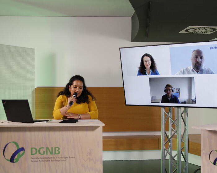 Zu sehen ist Seema Issar (DGNB) hinter einem Pult mit einem Mikrofon. Auf dem Monitor neben ihr sind zugeschaltet zu sehen: Sabine Döbeli, CEO, Swiss Sustainable Finance André Meyer, Leiter Kompetenzcenter Nachhaltige Immobilien, GLS Gemeinschaftsbank eG Sascha Rodenberg, Referent Nachhaltigkeit in der Wohnraumförderung, NRW.BANK